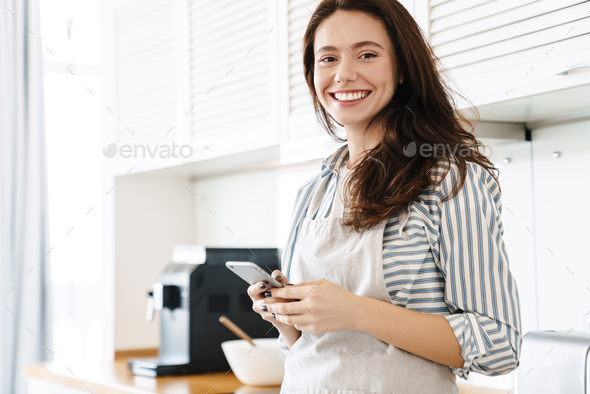 Image of smiling brunette woman using cellphone while cooking pie