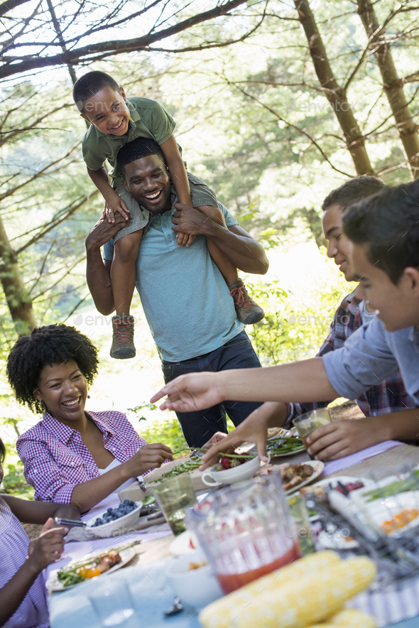 A family picnic meal in the shade of tall trees. A young boy sitting on his father\'s shoulders.