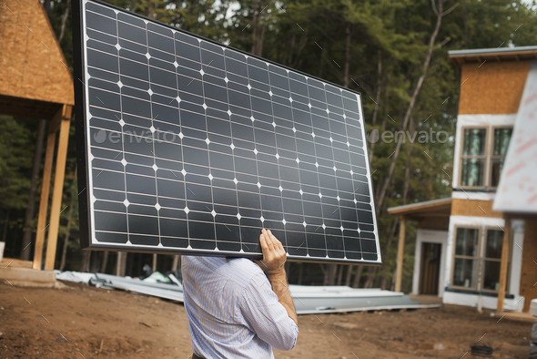 A workman carrying a large solar panel at a green house construction site.