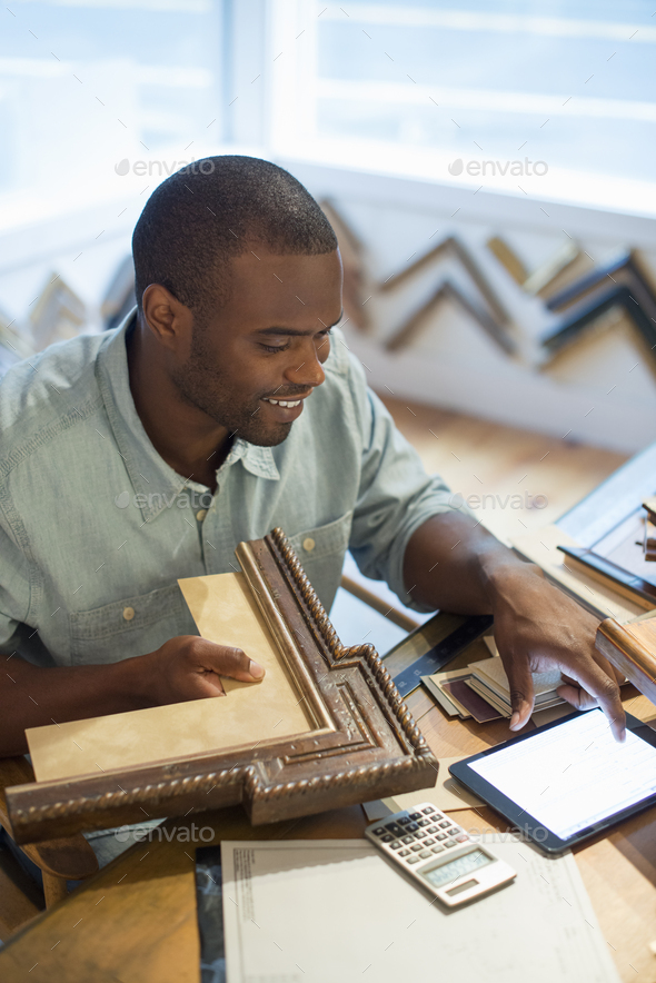 A young man at his workbench in a picture framing studio, using a digital tablet.