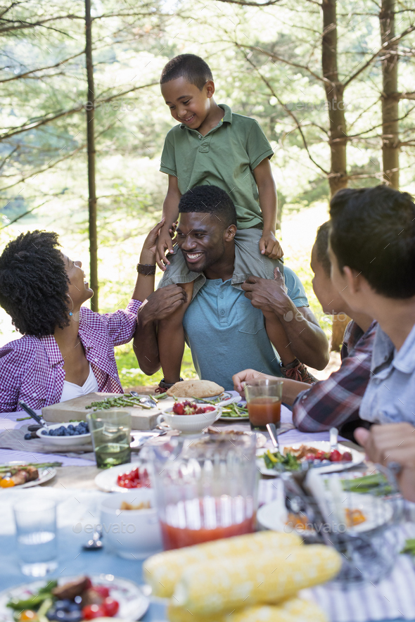 A family picnic meal in the shade of tall trees. A young boy sitting on his father\'s shoulders.