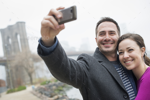 A couple taking a picture with a phone,a selfy of themselves by Brooklyn Bridge.