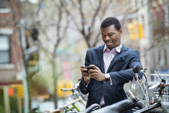 A young man in a blue suit,by a bicycle park. Checking his smart phone for messages.