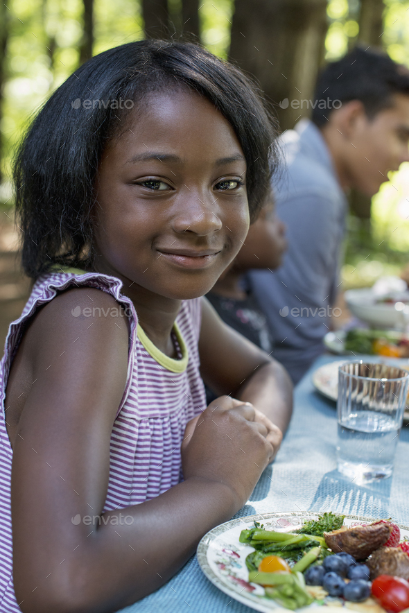 A family picnic meal in the shade of tall trees. A young girl seated at the table.