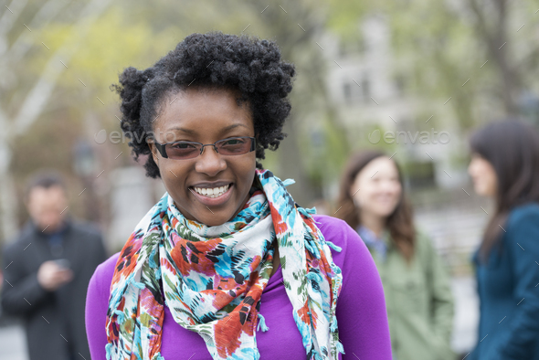 A group of people in a city park. A young woman smiling, wearing a purple shirt and floral scarf.