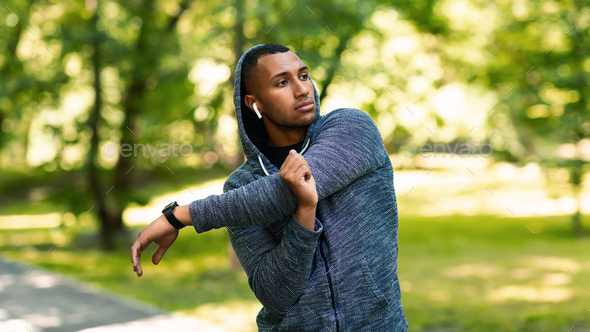 Athletic African American guy in hoodie stretching his arm and listening to music after training at