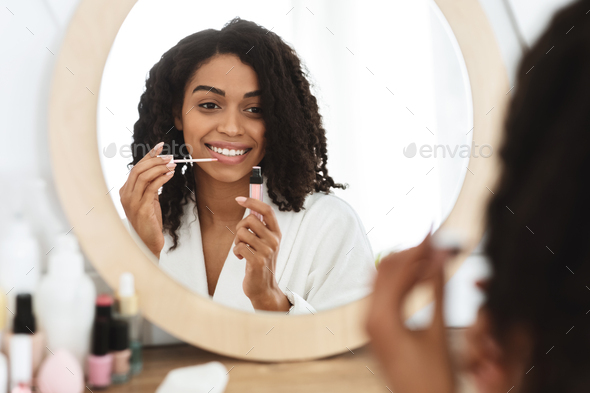Makeup Routine. Smiling African American Girl Applying Lip Gloss Near Mirror