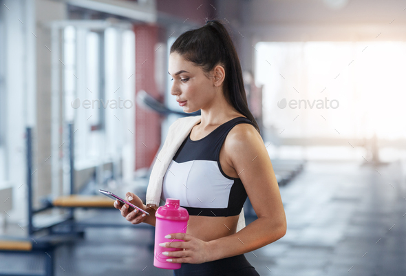 Millennial woman with mobile phone and bottle of water or protein shake at gym