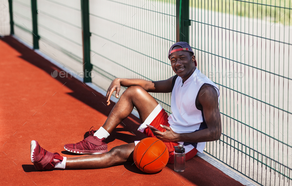 Handsome black sportsman with ball and mobile phone having rest at outdoor basketball court