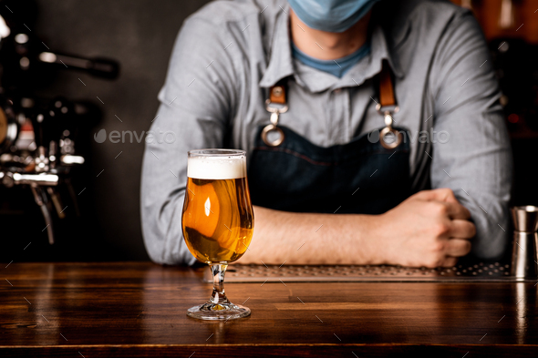 Bartender in protective mask leans on bar counter on which stands glass of light beer with foam