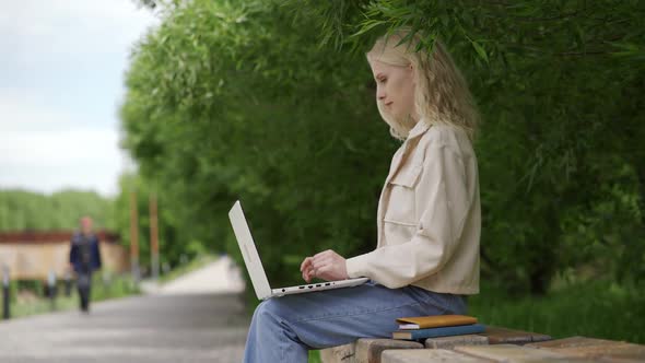 A Cute Student with Blonde Curls is Doing an Assignment Using a Laptop on a Park Bench
