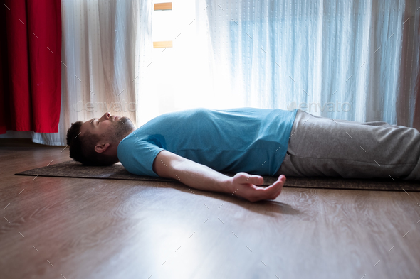 Young man meditating on a floor and lying in Shavasana pose at his living room. - Stock Photo - Images