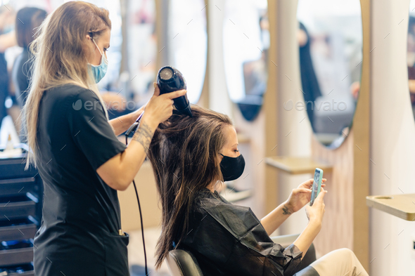 Hairdresser drying her client's hair with a hairdryer wearing protective masks in a beauty - Stock Photo - Images