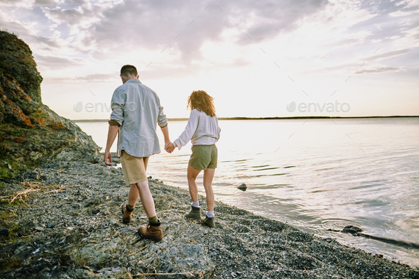 Young amorous hikers in casualwear holding by hands while moving along coastline