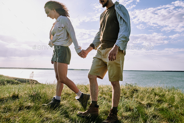 Relaxed young amorous hikers holding by hands while moving slowly down riverbank