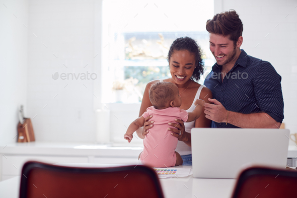 Family With Baby Daughter In Kitchen Using Laptop On Counter