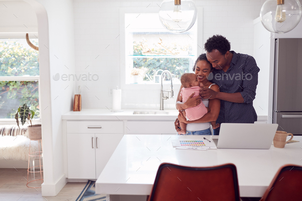 Family With Baby Daughter In Kitchen Using Laptop On Counter