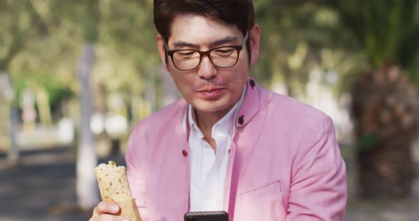 Asian man having a snack and using smartphone while sitting at the park alt