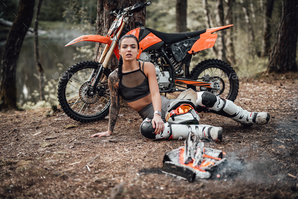 Young tattoed racer girl warming up while sitting next to the bonfire in the woods
