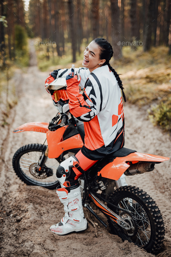 Happy female racer wearing motorcycle outfit sitting on her bike off-road  in the woods Stock Photo by fxquadro