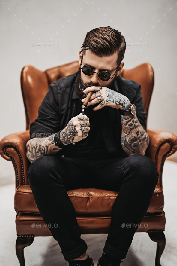 Bearded tattooed man with stylish haircut in sunglasses who smokes a cigar  Stock Photo by fxquadro