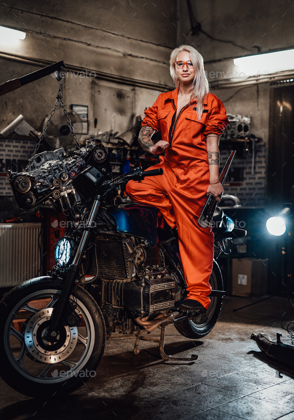 Beautiful female mechanic in orange overalls hold a big wrench and posing for a camera in garage - Stock Photo - Images