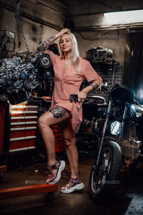 Beautiful blonde girl with tattooed body wearing pink dress posing in garage or workshop - Stock Photo - Images