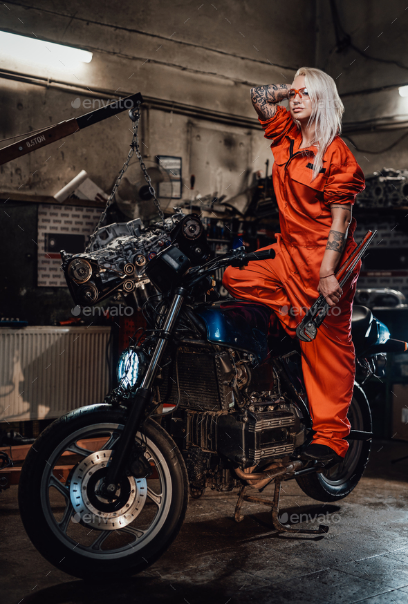Beautiful female mechanic in orange overalls hold a big wrench and posing for a camera in garage - Stock Photo - Images