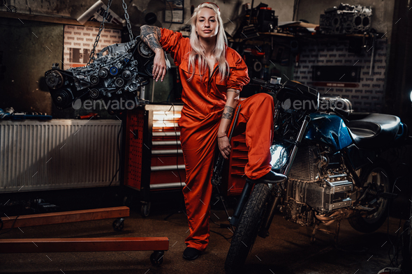 Blond female mechanic with tattooed hands in orange overalls stands in garage or workshop - Stock Photo - Images