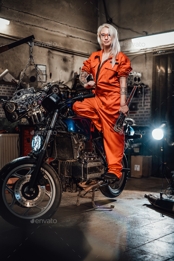 Beautiful female mechanic in orange overalls hold a big wrench and posing for a camera in garage - Stock Photo - Images