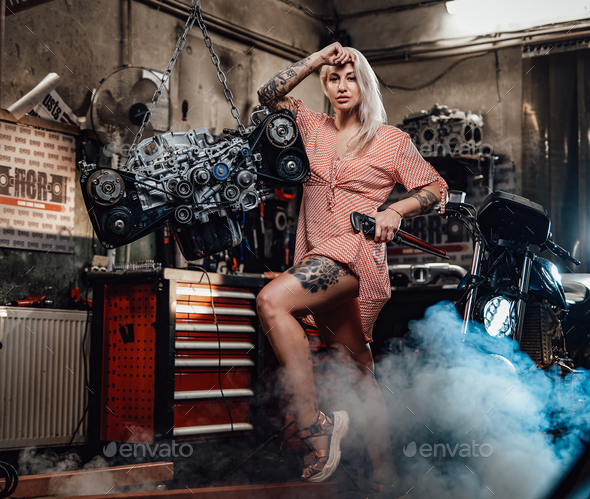 Beautiful blonde girl with tattooed body wearing pink dress posing in garage or workshop - Stock Photo - Images