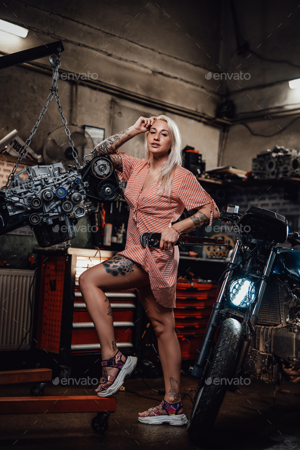 Beautiful blonde girl with tattooed body wearing pink dress posing in garage or workshop