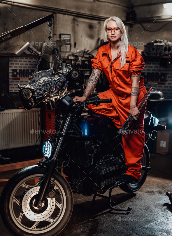 Beautiful female mechanic in orange overalls hold a big wrench and posing for a camera in garage - Stock Photo - Images