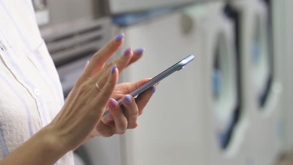 Closeup of a Woman's Hands with a Smartphone Against the Background of Washing Machines in a Public alt