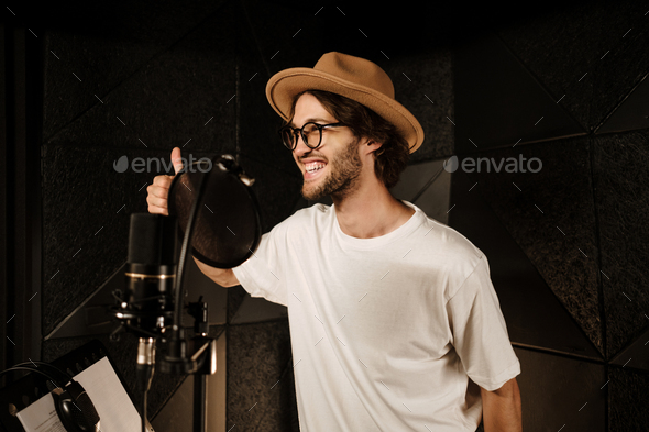 Attractive stylish male singer happily showing thumb up recording new song in modern sound studio