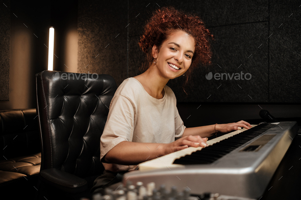 Young attractive woman happily looking in camera playing on electric piano in sound recording studio