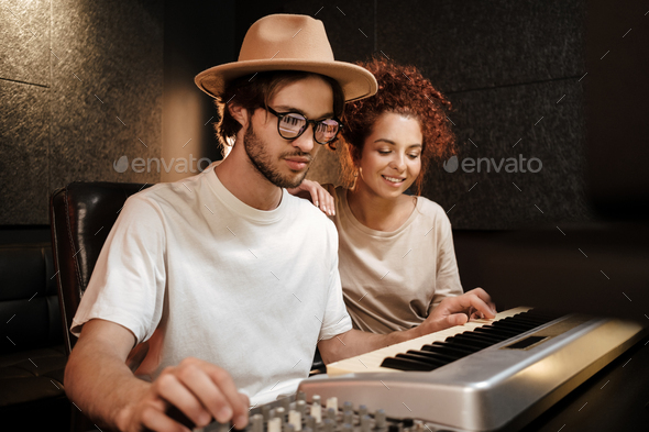Young stylish musicians happily recording new song on piano keyboard in modern sound studio