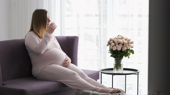 Pregnant Woman Stroking Her Big Belly While Relaxing on the Sofa at Home Near Bouquet of Rose