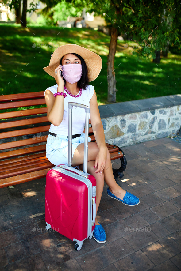 Woman protective mask, in the park outdoor with a suitcase, talking on a mobile - Stock Photo - Images