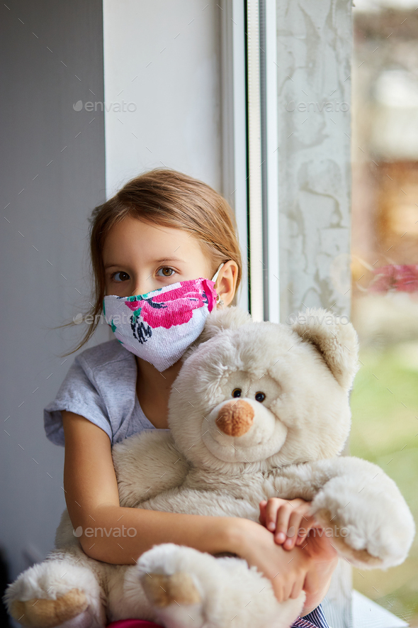 Little girl, child in mask with teddy bear sits on windows, coronavirus quarantine - Stock Photo - Images