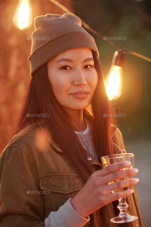 Happy Asian girl with dark long hair toasting with drink against lights - Stock Photo - Images