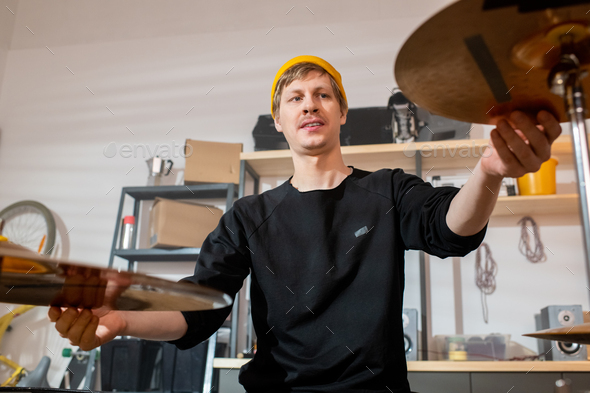 Young musician in black pullover and yellow beanie touching cymbals of drum kit