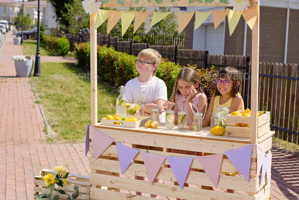 Group of adorable girls and boy standing by wooden stall and selling lemonade