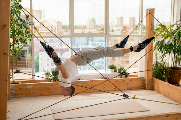 Young sportsman with his arms and legs fixed to ropes doing aerial yoga exercise