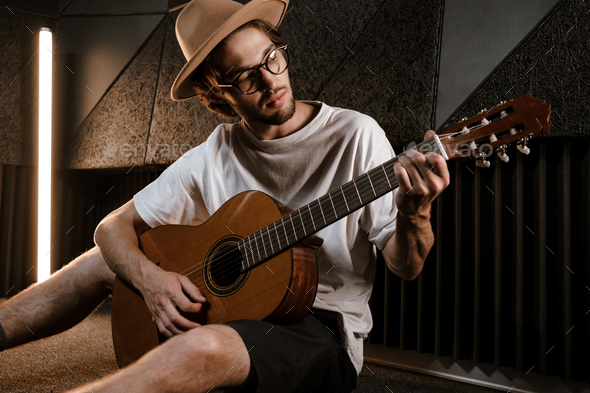 Young attractive male musician thoughtfully playing on guitar in sound recording studio