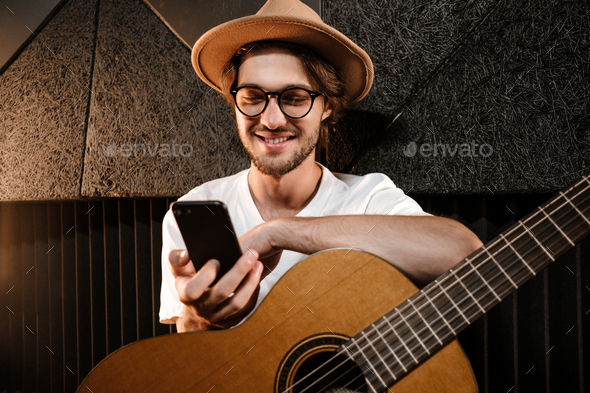 Young attractive man with guitar happily using smartphone in modern sound recording studio