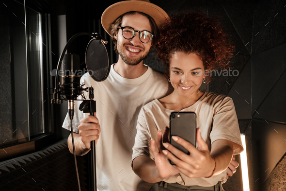 Couple of attractive singers happily taking selfie together on smartphone in sound recording studio