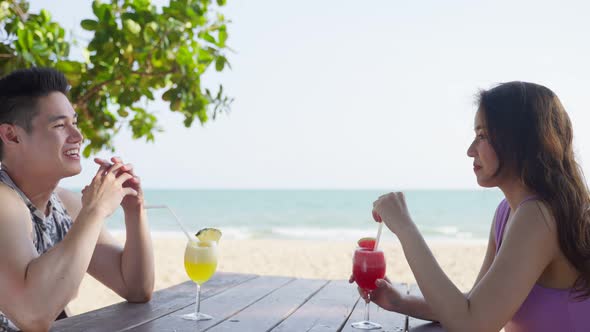 Asian young man and woman couple drinking juice on the beach together on holiday vacation. alt