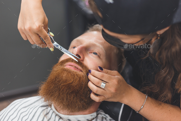 A man at a barbershop. Woman barber clipping mustache. Barber woman in mask.