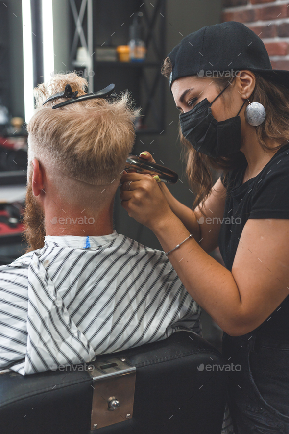 Female barber in mask cuts a man hairs with hair clipper. Hairstyle during social distancing